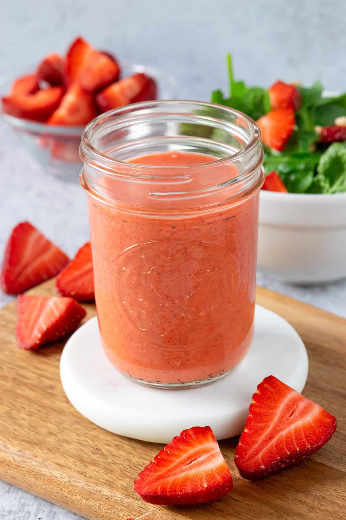 A jar of homemade strawberry vinaigrette surrounded by a few cut strawberries. In the background there's a bowl of strawberries and a strawberry spinach salad.