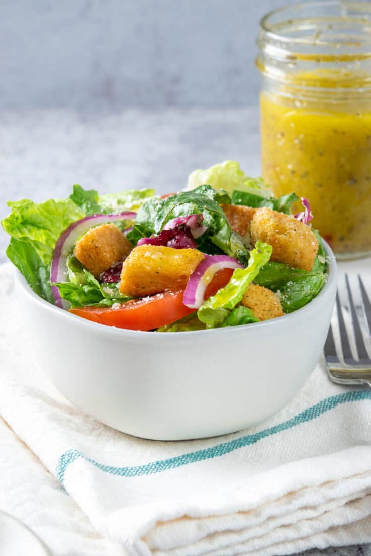 A green alad in a bowl with croutons, tomatoes and red onion that's drizzled in Italina dressing. A small mason jar filled with the homemade vinaigrette is in the background.