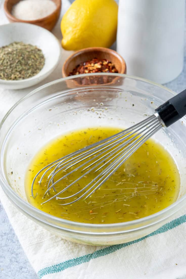 A bowl of homemade Italian vinaigrette that has a whisk in it. The bowl is in front of a bowls of red pepper flakes, garlic powder and Italian seasoning, and a lemon, and bottle of olive oil.