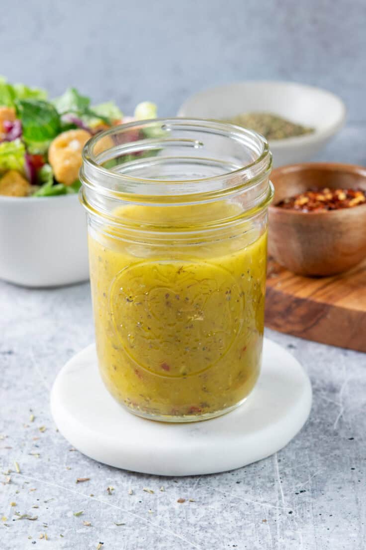 Homemade Italian dressing in a pint mason jar that's next to a bowl of red pepper flakes, a bowl of Italian seasoning and a salad that's topped with croutons.