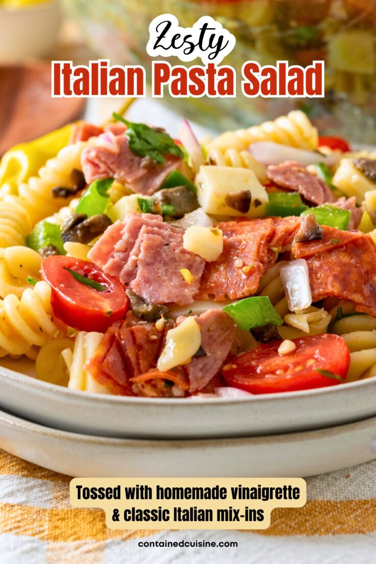 Homemade Italian pasta salad piled on a plate. The close-up photo shows rotini pasta, covered with Italian dressing, with salami, pepperoni, provolone cheese, pepperoncini, olives and veggies mixed in. The image has a text overlay heading that says, zesty Italian pasta salad, and a sub-heading that says, tossed with homemade vinaigrette and classic Italian mix-ins.