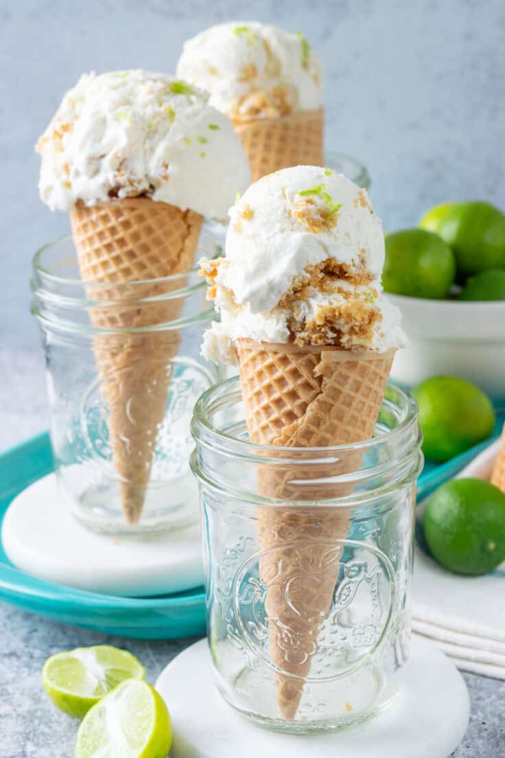 Homemade key lime pie ice cream scooped into three cones. The scoops of ice cream are filled with buttery graham cracker crust pieces and you can see flecks of lime zest in the ice cream. The cones are standing up in mason jar glasses and are surround by key limes.