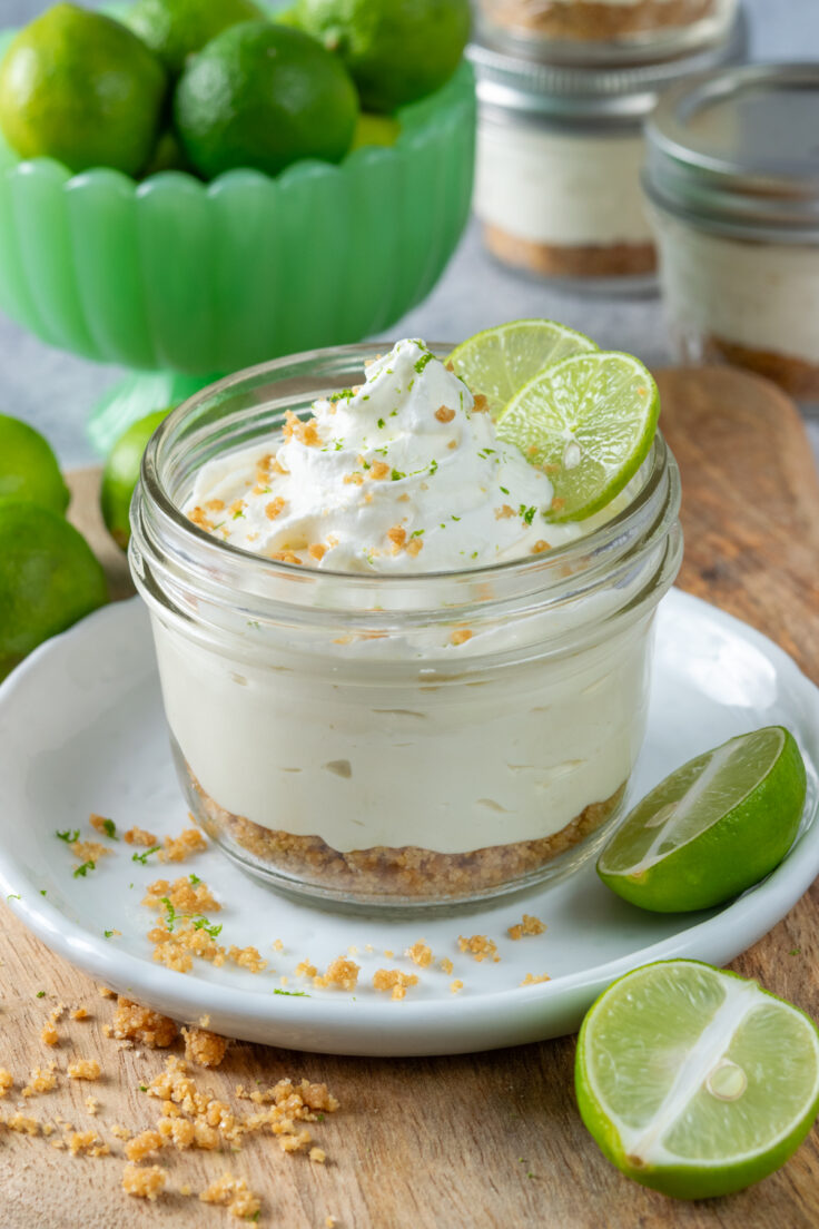 A single serving of key lime pie in a mason jar. The mini dessert has a layer of graham crackers on the bottom, creamy key lime filling in the middle, and whipped cream on top. The jar dessert is garnished with a slice of lime. In the background is a bowl of key limes and more jars of key lime pie.