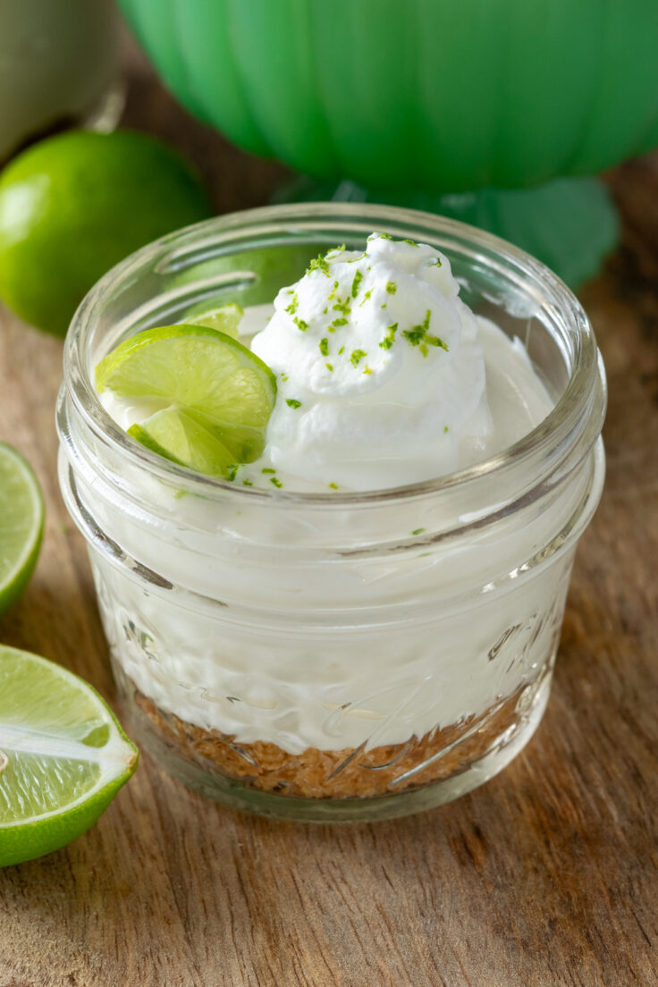 A single serving of key lime pie in a mini 4-ounce mason jar. The dessert is topped with a dollop of whipped cream, lime zest and a twisted slice of lime.