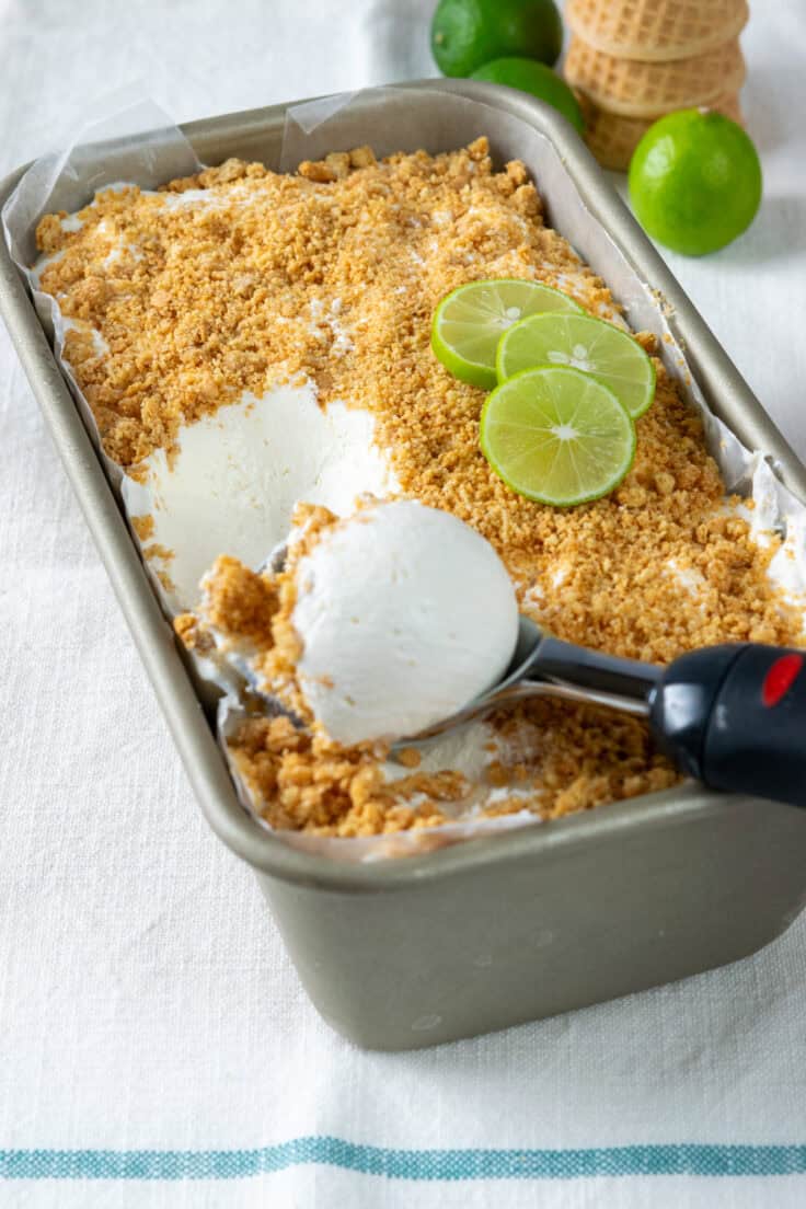 Homemade key lime pie ice cream frozen in a loaf pan. The ice cream has a buttery layer of graham crackers on top and layered into the  ice cream. There's an ice cream scoop on top which has scooped out some of the homemade ice cream. In the background are some key limes and a stack of ice cream cones.
