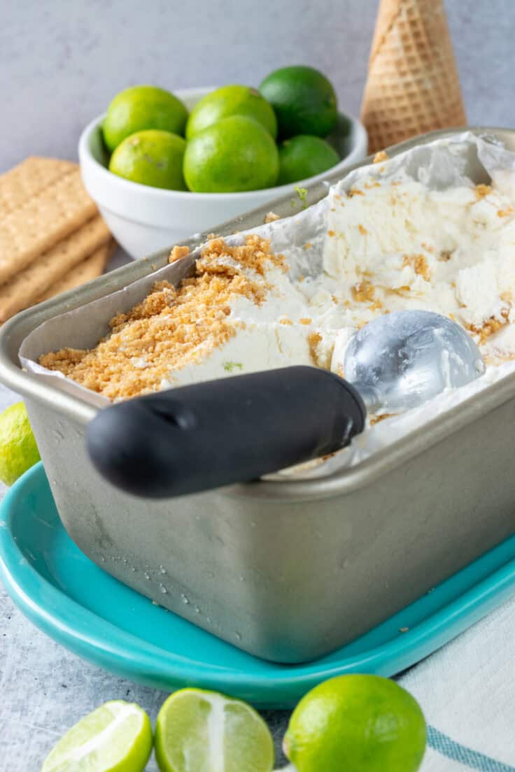 Homemade, no-churn key lime pie ice cream that's been frozen in a loaf pan. There's an ice cream scoop on top of the ice cream which has had a lot scooped out of it. Behind the ice cream is a stack of graham crackers, a small bowl of key limes, and some sugar cones.