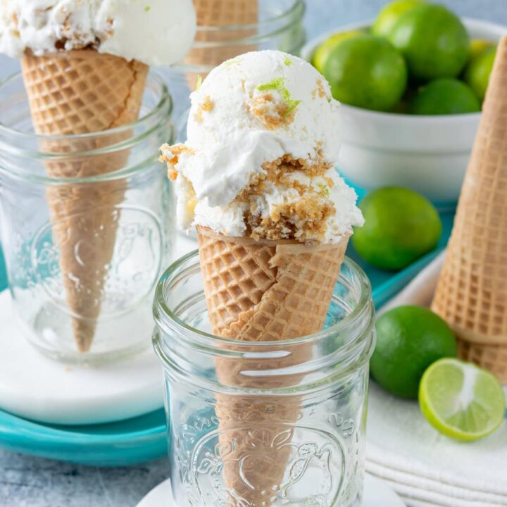 Homemade no-churn key lime pie ice cream is scooped into three ice cream cones. The cones are each standing in a mason jar. There are key lime halves and a bowl of key limes in the foreground and background.
