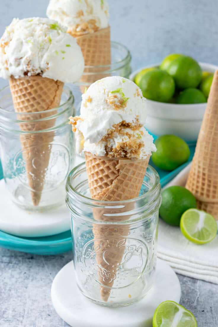 Homemade no-churn key lime pie ice cream is scooped into three ice cream cones. The cones are each standing in a mason jar. There are key lime halves and a bowl of key limes in the foreground and background.