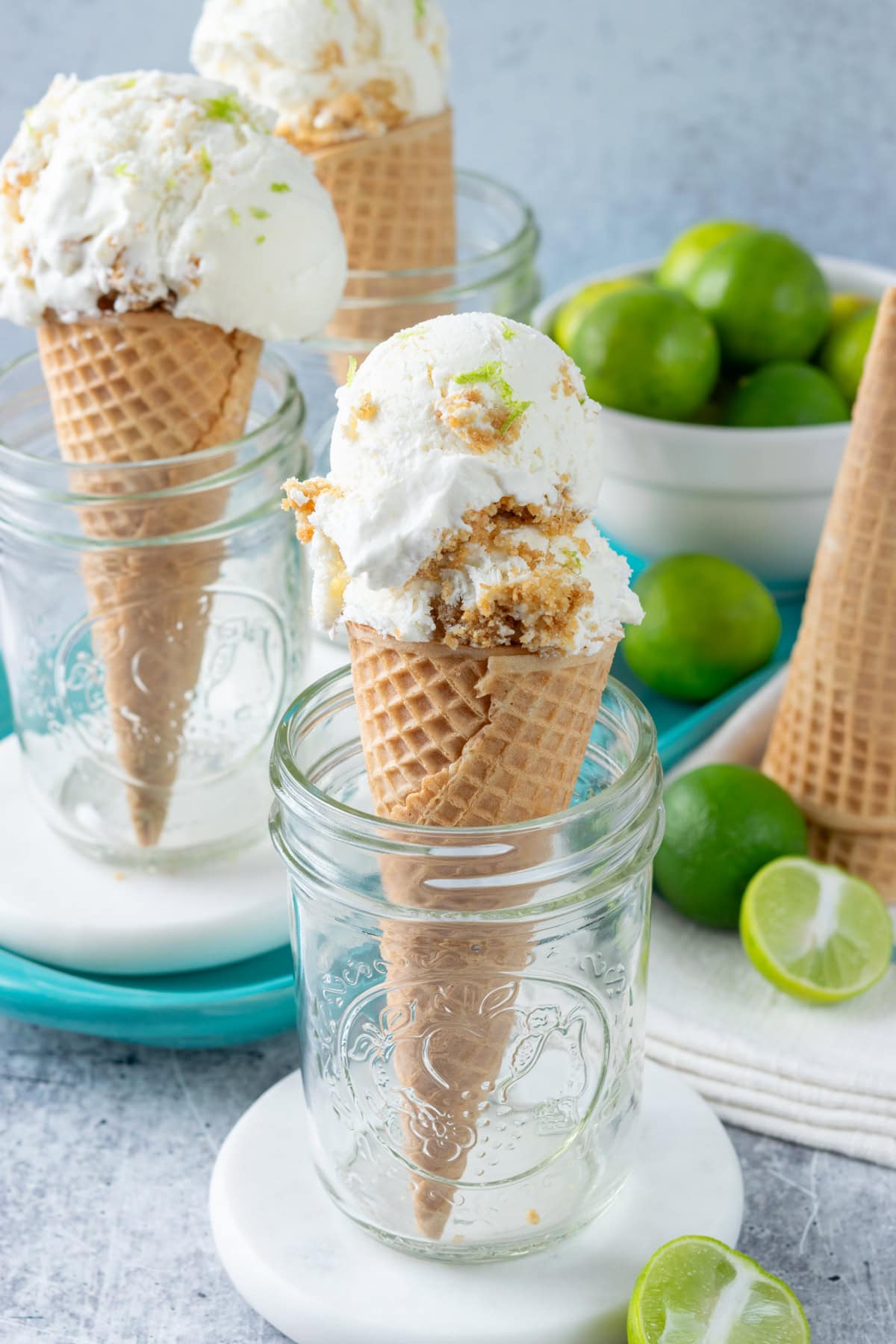 Homemade no-churn key lime pie ice cream is scooped into three ice cream cones. The cones are each standing in a mason jar. There are key lime halves and a bowl of key limes in the foreground and background.