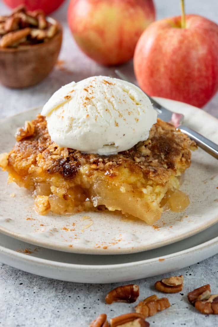 A warm and gooey slice of apple dump cake with ice cream melting on top. The dessert is next to a few red apples and a small wooden bowl filled with pecans.