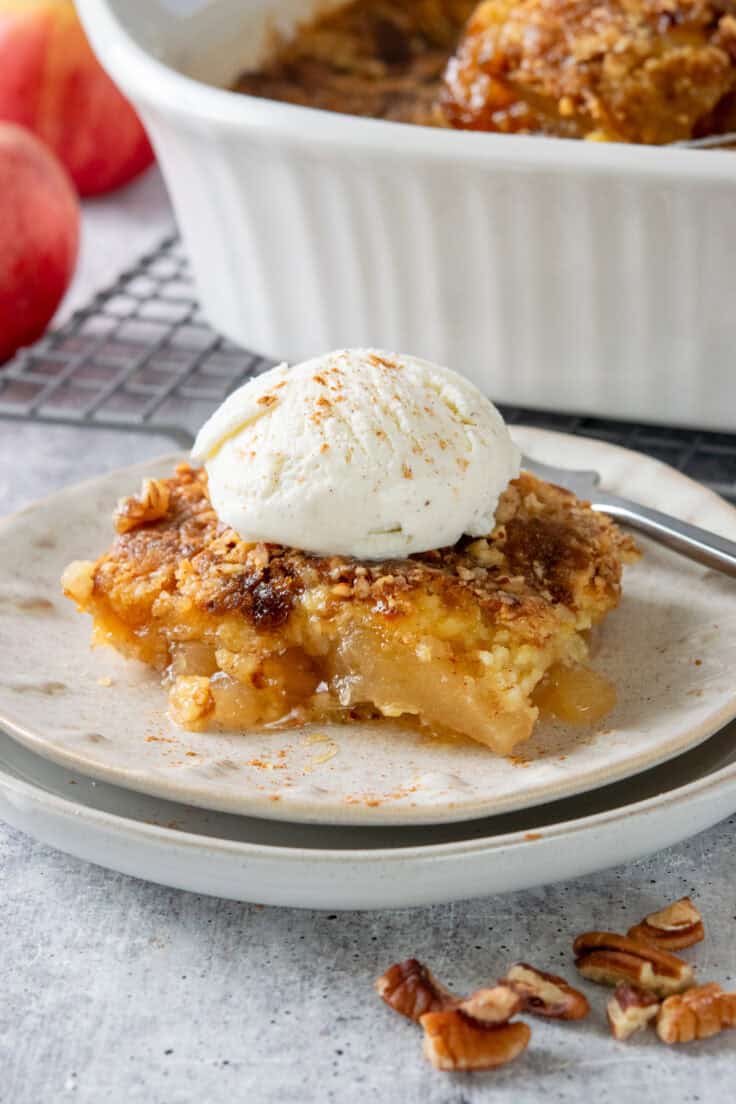 A serving of apple dump cake and ice cream on a plate next to the baking dish full of the dessert. There are also a couple of red apples.