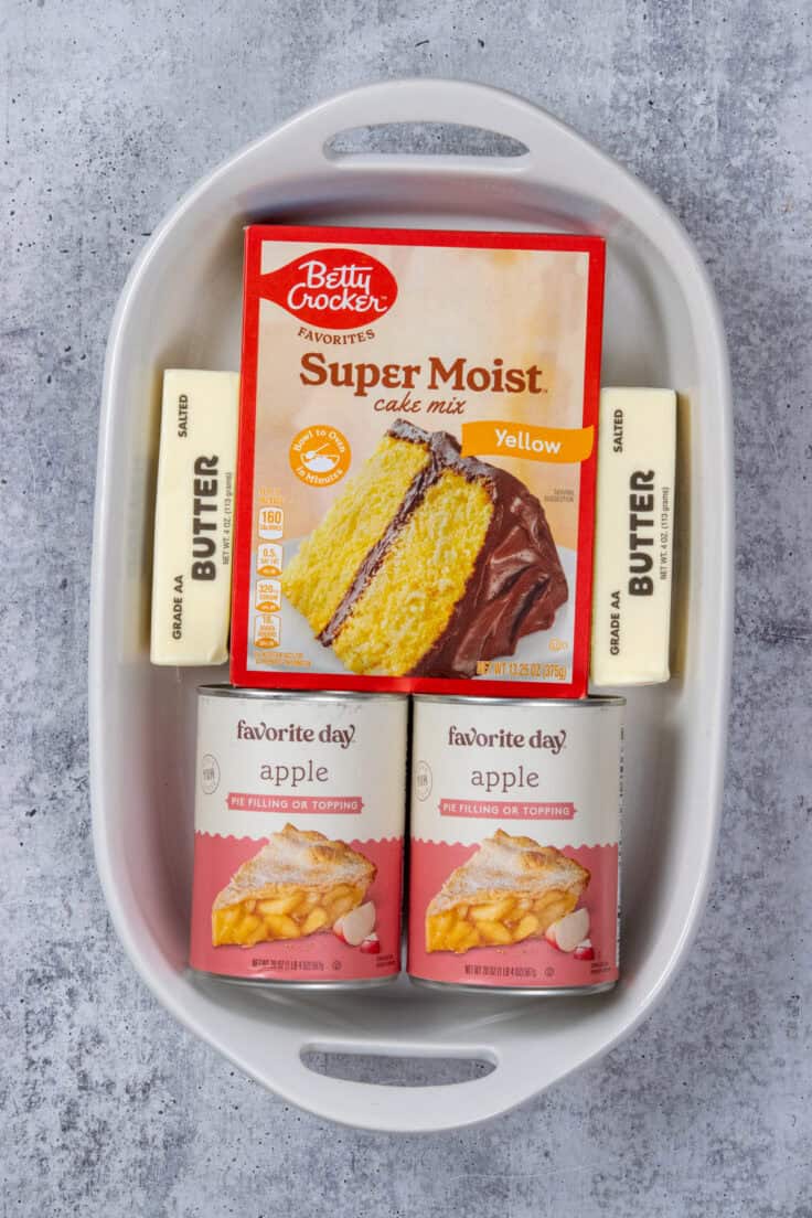 A 9x13 baking that has the three main ingredients in it. There's a boxed yellow cake mix, two cans of apple pie filling, and two sticks of salted butter.