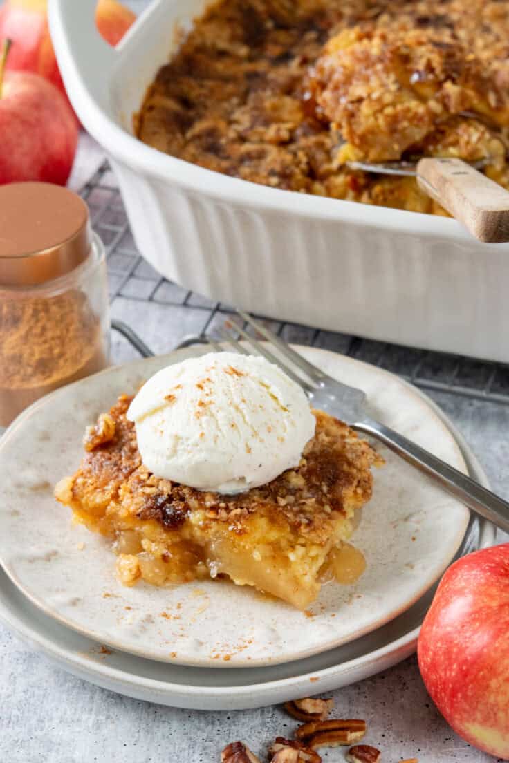 A serving of apple dump cake with a scoop of ice cream on top. It's next to baking dish full of the warm, apple cobbler-like dessert.