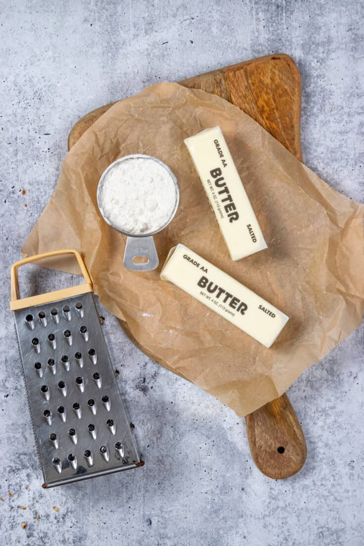 Two sticks of butter and a small amount of cake mix on a piece of parchment paper next to a box grater.