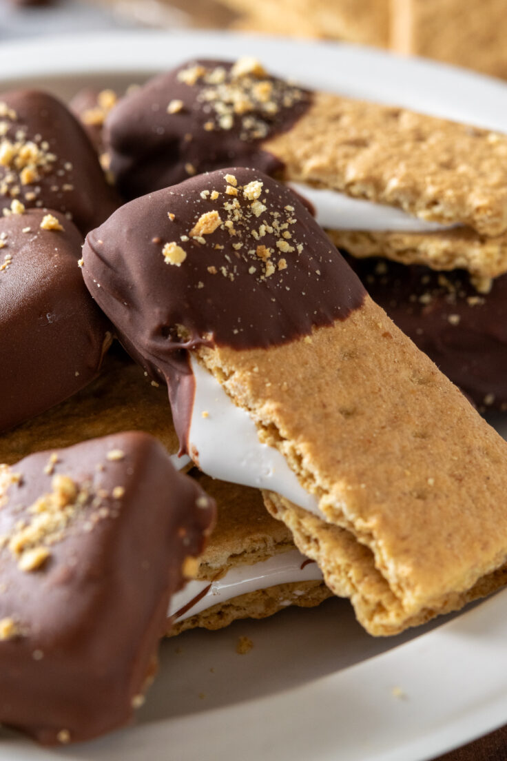 This image shows a close-up of a graham cracker smores bite. Marshmallow fluff is stuffed between two graham crackers and dripping out. One end of the treat is dipped in milk chocolate and dusted in graham cracker crumbs.