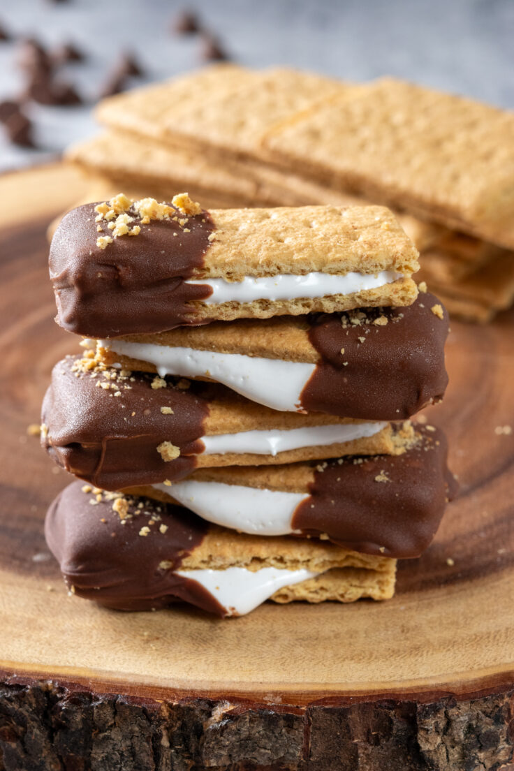 Four s'mores bites stacked on top of each other. You can see a thick layer of marshmallow fluff sandwiched between the graham crackers. One end of the bites is dipped in milk chcolate and decorated with graham cracker crumbs.