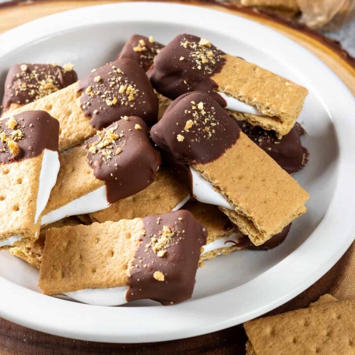 A plate piled high with marshmallow fluff smores bites. Marshmallow fluff is oozing out of each graham cracker bite which is dipped in chocolate.