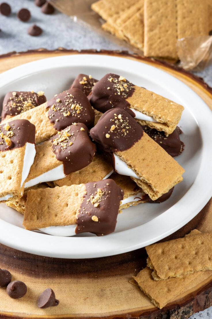 A plate piled high with marshmallow fluff smores bites. Marshmallow fluff is oozing out of each graham cracker bite which is dipped in chocolate.