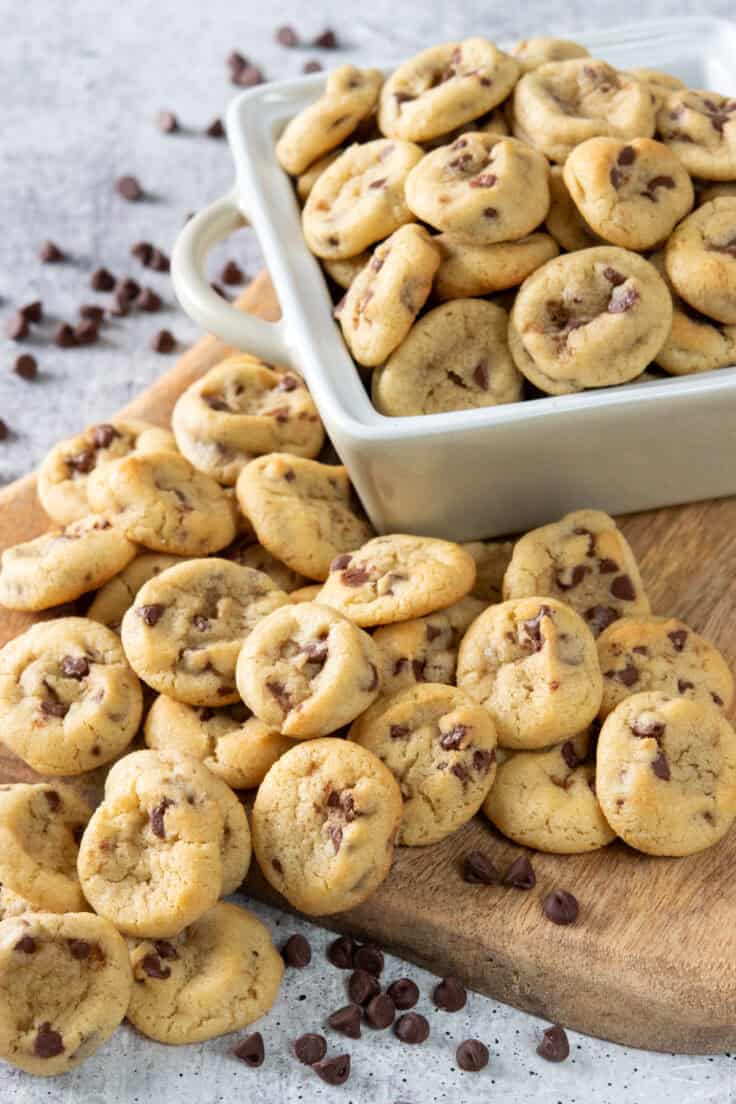 Piles of homemade mini chocolate chip cookies on a wooden board and in a small baking dish, surrounded by mini chocolate chips scattered around.