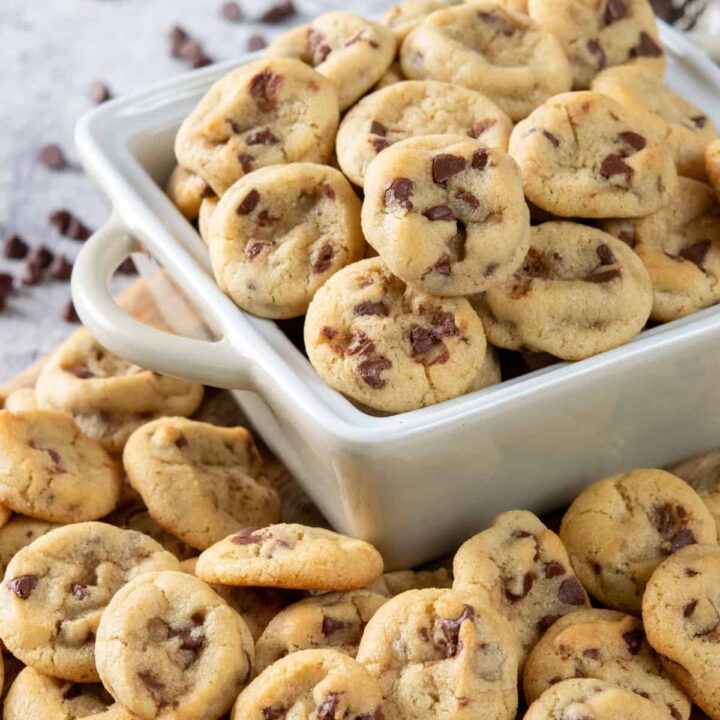 A small dish filled with homemade mini chocolate chip cookies. There are a bunch of tiny cookies surrounding the dish and some mini chocolate chip morsels scattered in the background.
