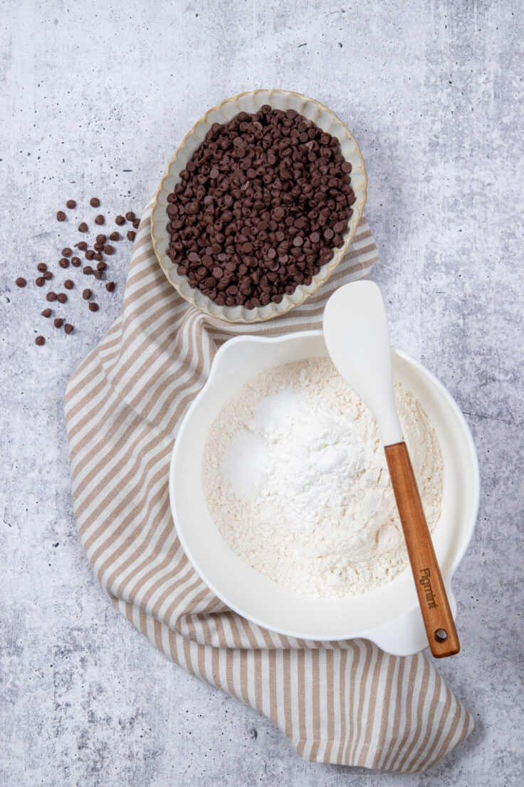 A mixing bowl filled with flour, baking soda, salt and corn starch to make cookie dough. There's a rubber spatula resting on the mixing bowl and a small bowl filled with mini chocolate chips.