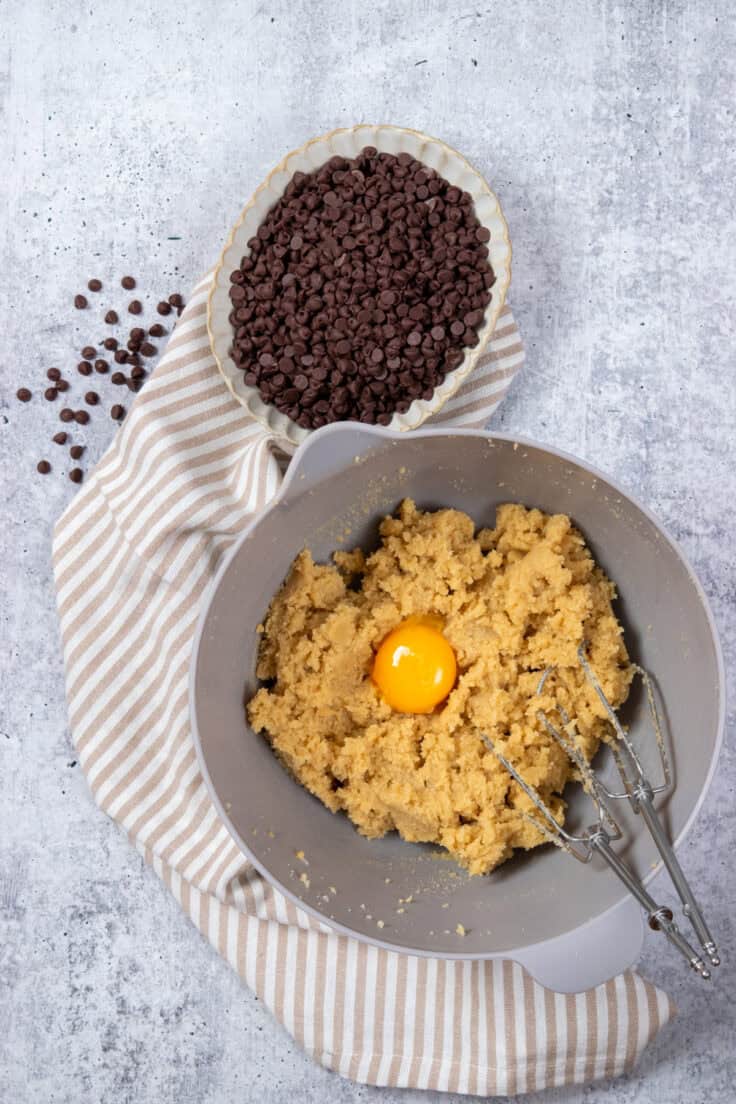 A mixing bowl showing an egg being beaten into the butter, sugar and vanilla cookie dough batter. There's a small bowl of mini chocolate chips next to the mixing bowl.