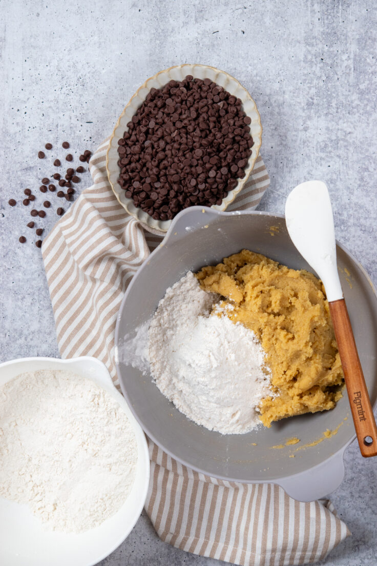 Half of the dry mixture being stirred into the bowl of wet batter to make the cookie dough. There's a bowl of mini chocolate chips next to the mixing bowl.