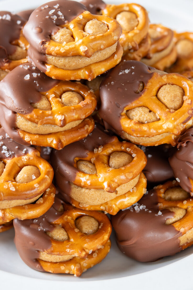 A close-up photo of homemade pretzel bites stacked on a platter. You can see the creamy peanut butter filling between the pretzels and how half of each treat is dipped in chocolate and finished with a touch of sea salt.