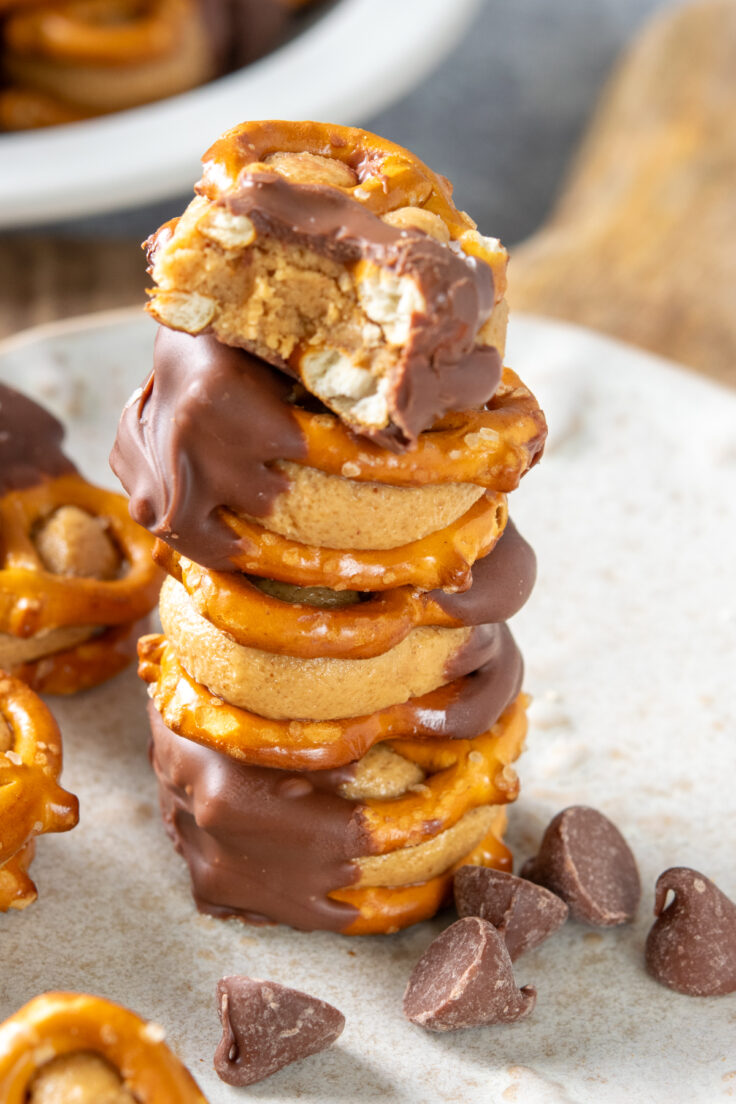 A stack of peanut butte pretzel bites  surrounded by loose chocolate chips. The top treat has a bite out of it showing the creamy and crunchy inside.
