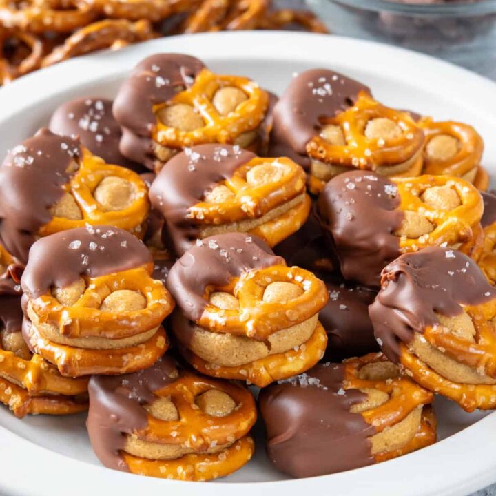 Homemade peanut butter pretzel bites piled up on a plate. Each treat has two pretzels with sweetened peanut butter balls flattened in-between, and then half is dipped in chocolate with flaky sea salt sprinkled on top.