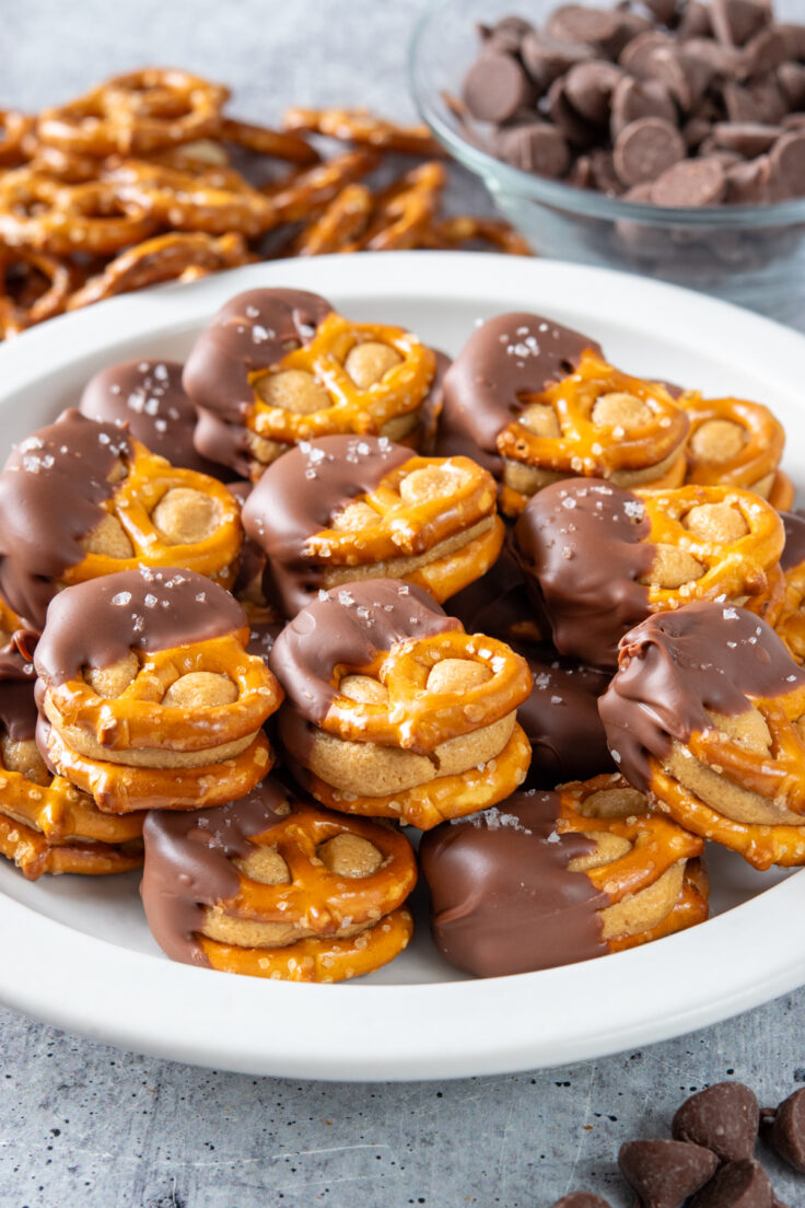Homemade peanut butter pretzel bites piled up on a plate. Each treat has two pretzels with sweetened peanut butter balls flattened in-between, and then half is dipped in chocolate with flaky sea salt sprinkled on top.