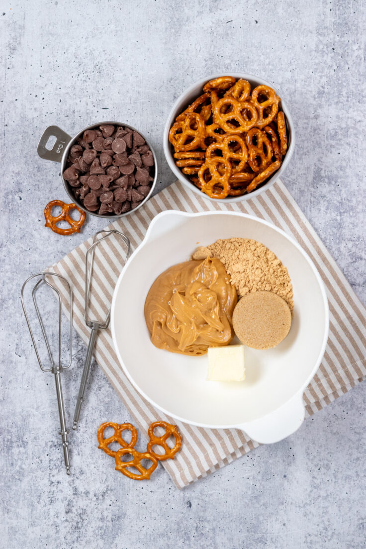 A mixing bowl containing peanut butter, powdered peanut butter, brown sugar and softened butter. Next to the bowl are some beaters and bowls with chocolate chips and mini pretzels.