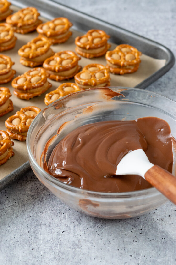 A bowl of melted chocolate next to a tray of 36 peanut butter pretzel bites waiting to be dipped in chocolate.