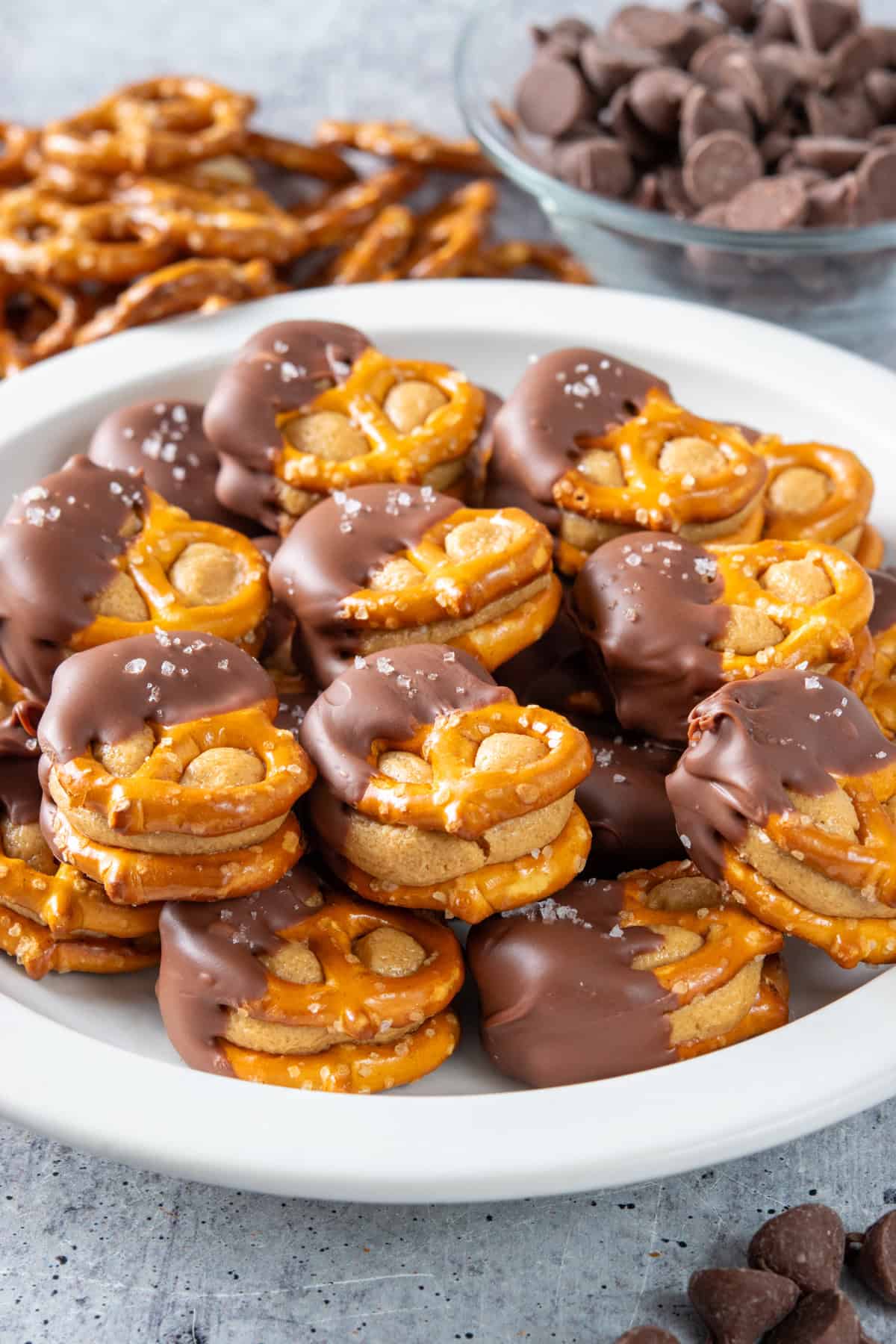Homemade peanut butter pretzel bites piled up on a plate. Each treat has two pretzels with sweetened peanut butter balls flattened in-between, and then half is dipped in chocolate with flaky sea salt sprinkled on top.