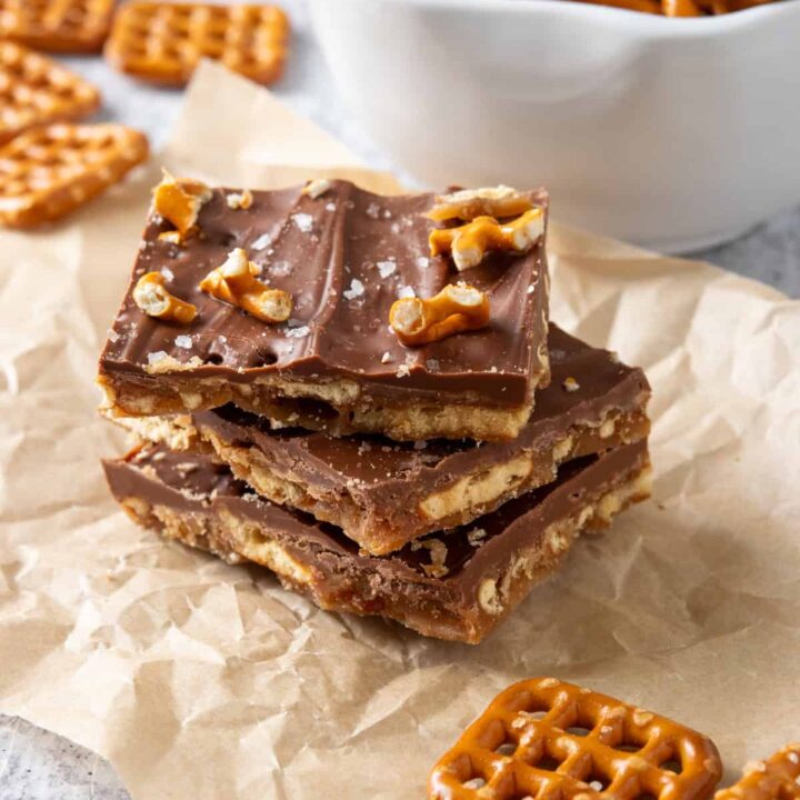 A stack of three pieces of salted caramel pretzel bark on a piece of parchment paper surrounded by some pretzels.