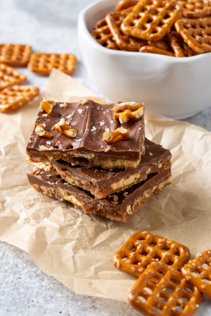 A stack of three pieces of salted caramel pretzel bark on a piece of parchment paper surrounded by some pretzels.