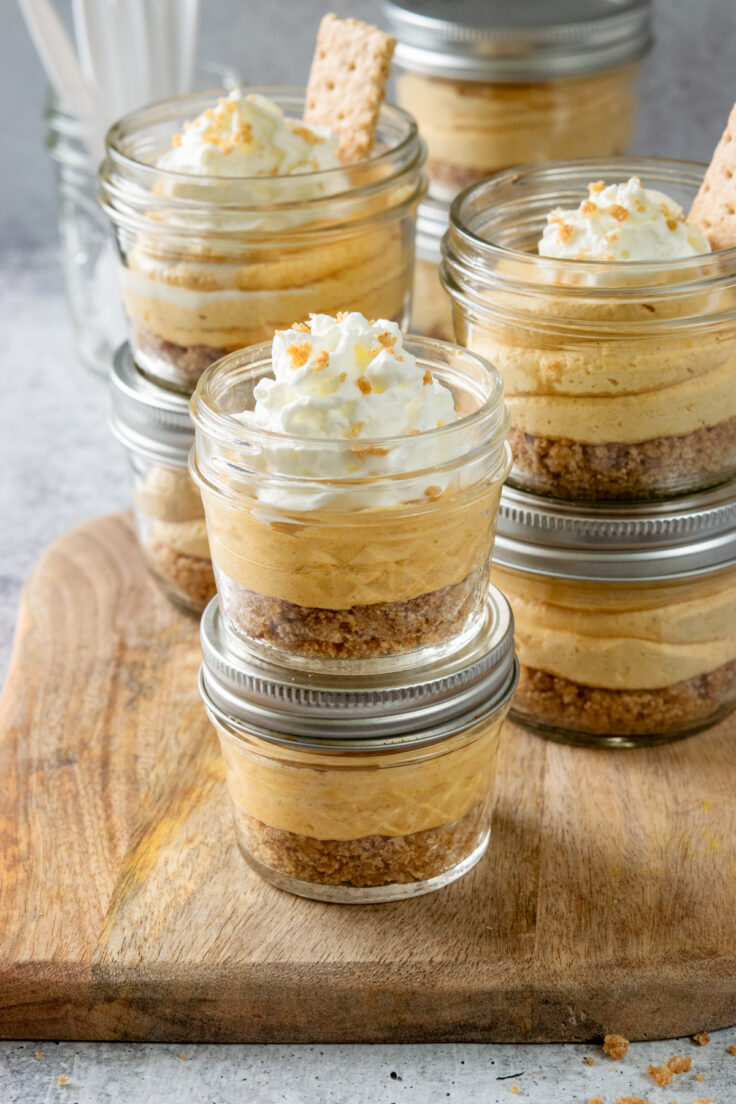 Two mini 4-ounce pumpkin cheesecake cups in small mason jars, in front of lots of 8-ounce pumpkin cheesecake jars. The pumpkin dessert is topped with whipped cream and some graham cracker crumbs. There's also a small cracker garnishing each dessert.