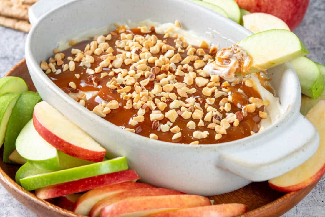 Gooey caramel apple cheesecake dip in a small dish which is on a tray with lots of red and green sliced apple. There are also graham crackers near the tray. In the dip is a piece of apple that's just been dipped and has lots of swirled together sweet cream cheese, caramel and nuts.