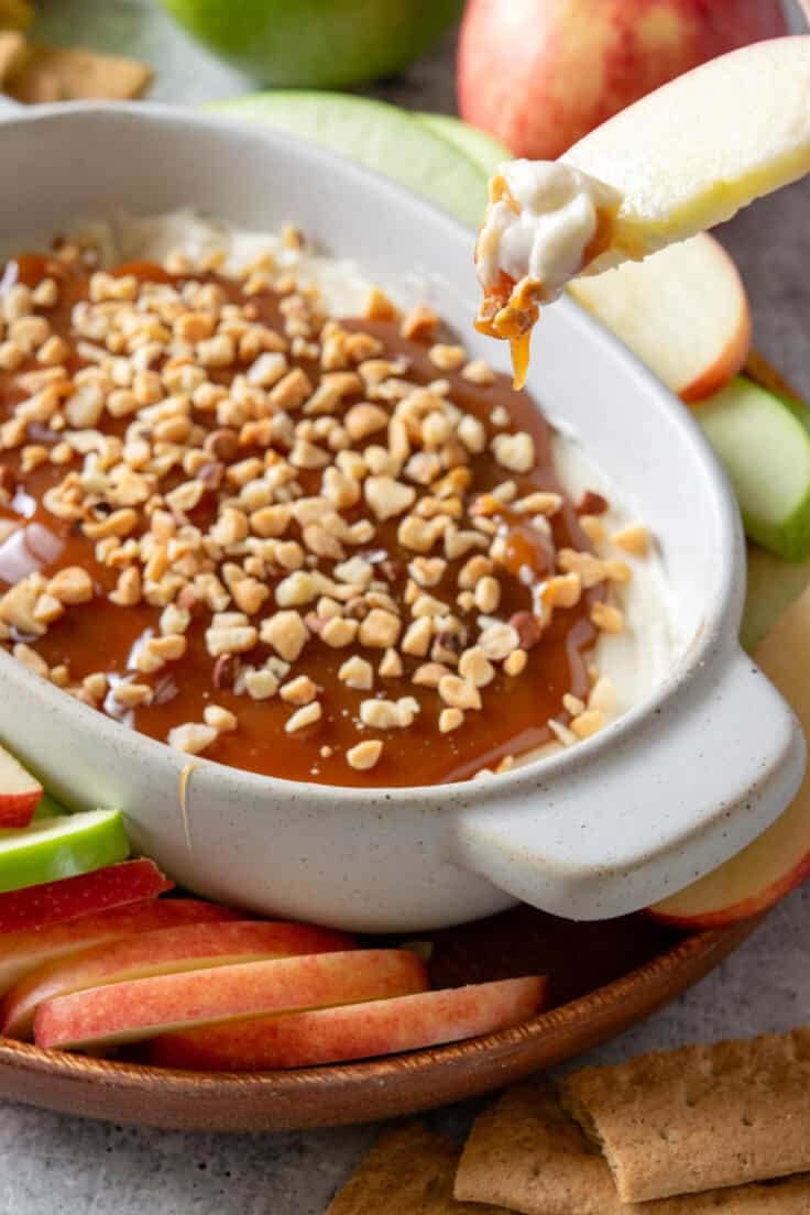 An apple slice that's been dipped in the dish of caramel cheesecake dip. There is caramel dripping off the apple.