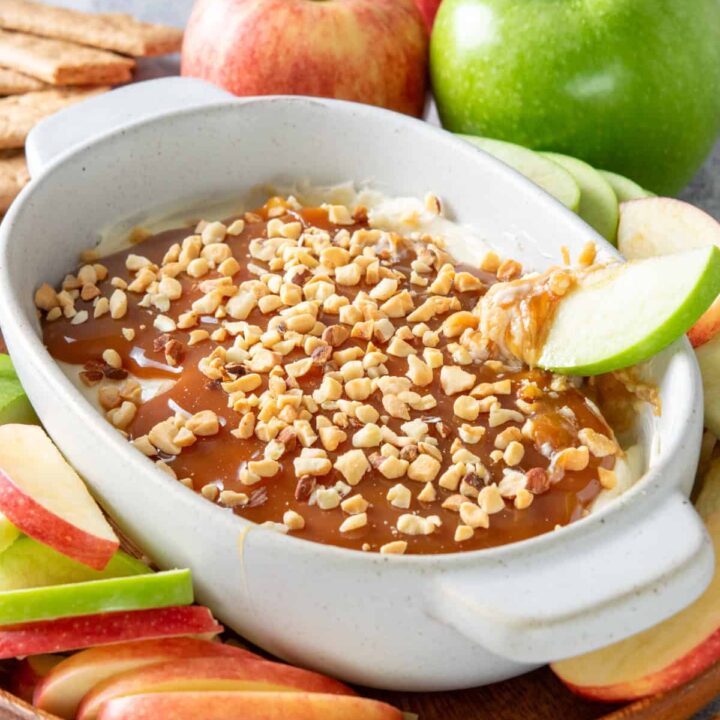 Caramel cheesecake dip with a granny smith apple slice being dipped into it. The apple has caramel, nuts and sweet cream cheese on it. The dessert dip is on a platter that has apple slices and graham crackers surrounding it.