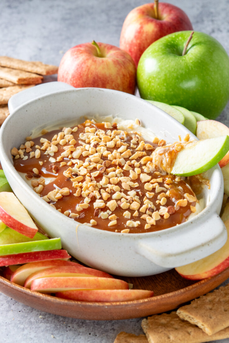 Caramel cheesecake dip with a granny smith apple slice being dipped into it. The apple has caramel, nuts and sweet cream cheese on it. The dessert dip is on a platter that has apple slices and graham crackers surrounding it.