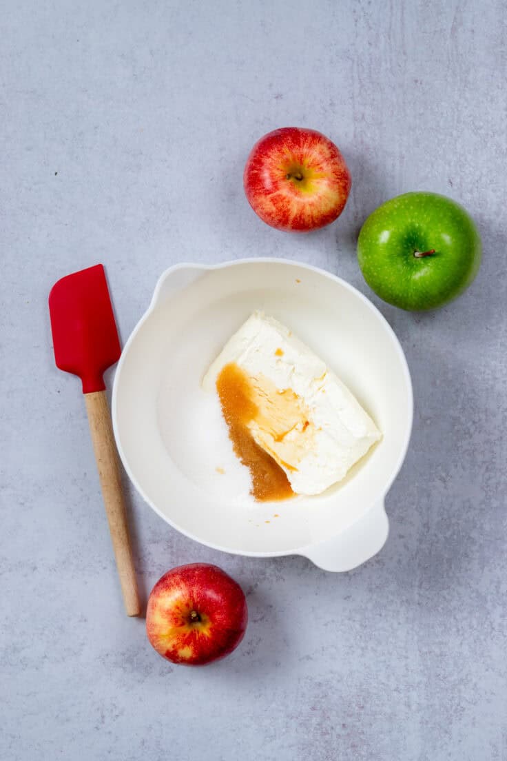 A mixing bowl with a block of cream cheese, sugar and vanilla. There are three apples and a red rubber spatula next to the bowl.