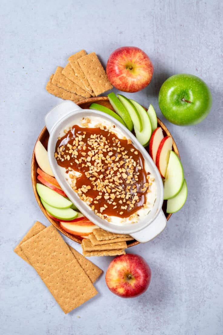 The caramel dip is on a tray which has a variety of red and green sliced apples and graham crackers on it for dipping.