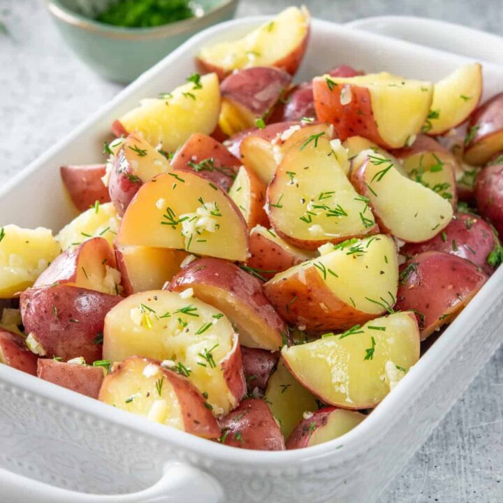 Buttery garlic dill red potatoes in a serving dish. There is fresh minced dill and a bulb of garlic behind the side dish.