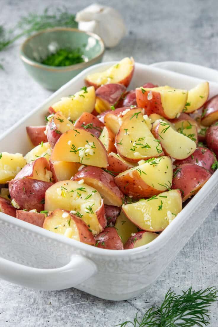 Buttery garlic dill red potatoes in a serving dish. There is fresh minced dill and a bulb of garlic behind the side dish.