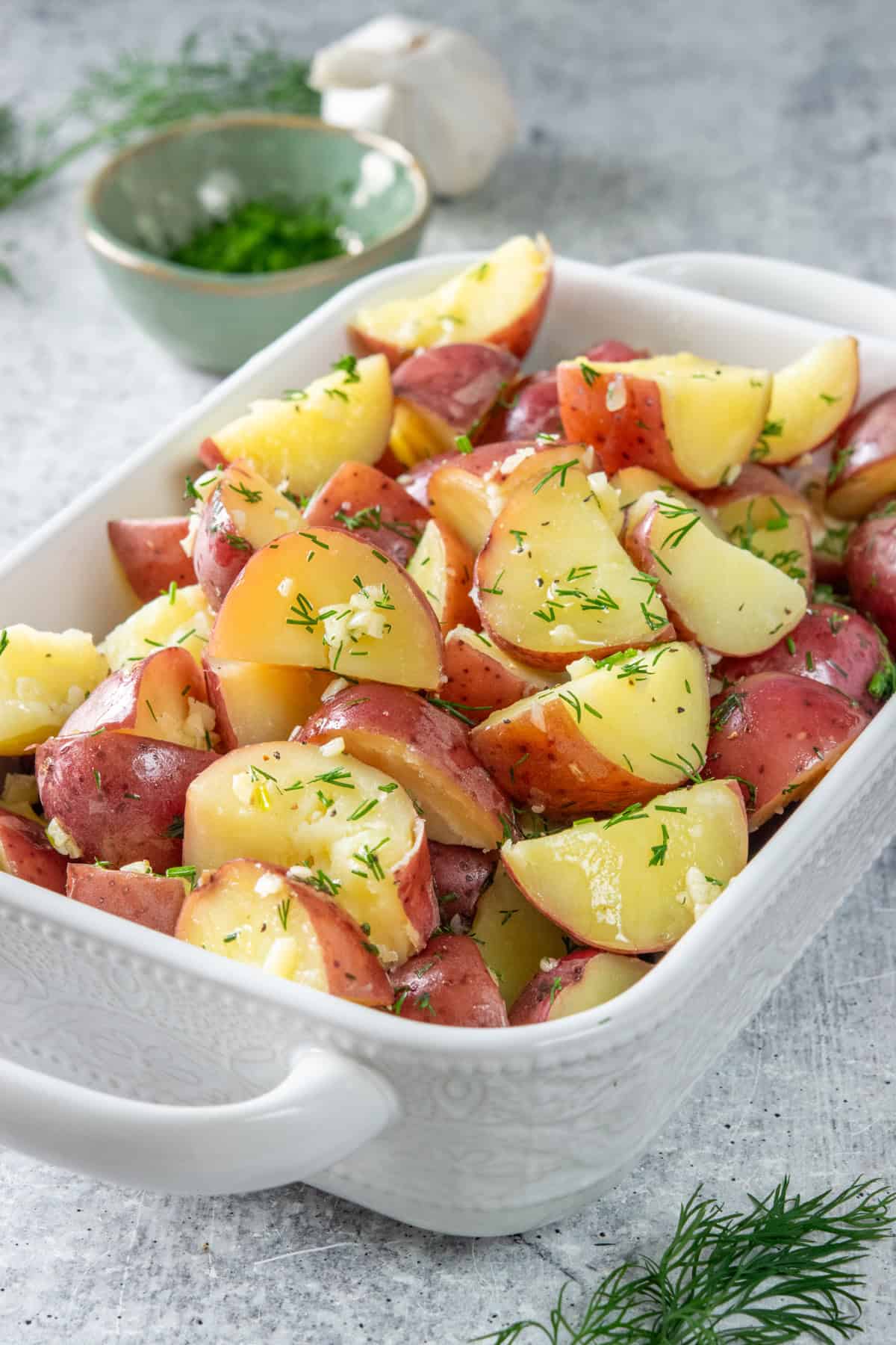 Buttery garlic dill red potatoes in a serving dish. There is fresh minced dill and a bulb of garlic behind the side dish.