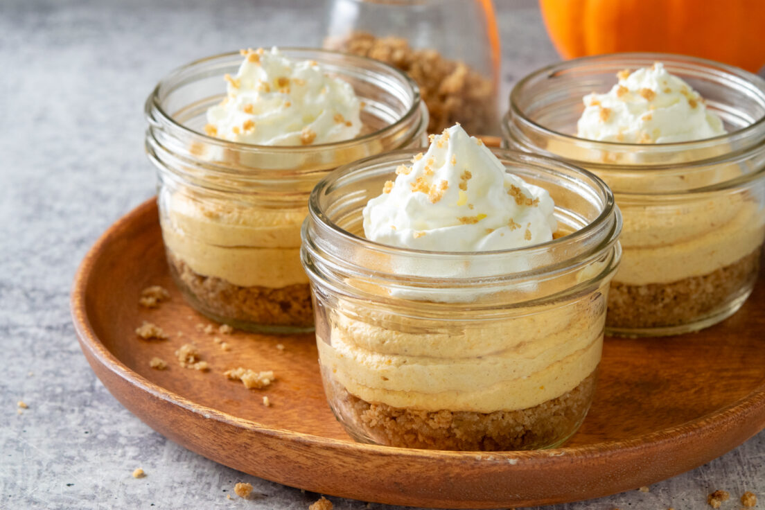 Three mini pumpkin cheesecake cups on a serving tray which is next to a pumpkin and jar of extra buttey graham cracker crumbs for spinkling on top of the dessert.