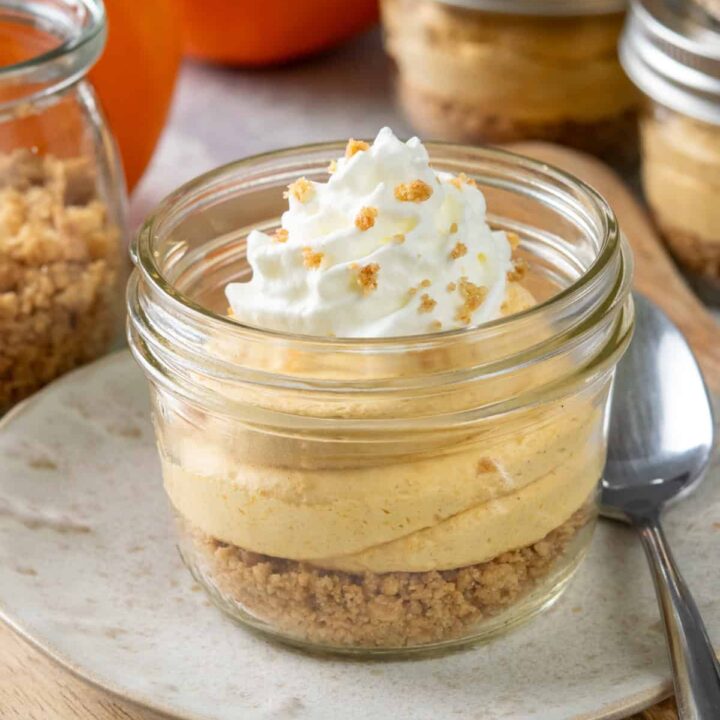 A pumpkin cheesecake cup that has a layer of graham cracker crumb crust, pumpkin cream cheese filling, and swirl of whipped cream. Behind the dessert cup are more pumpkin cups, a jar of buttery graham cracker crumbs to sprinkle on top of the dessert, and a pumpkin.