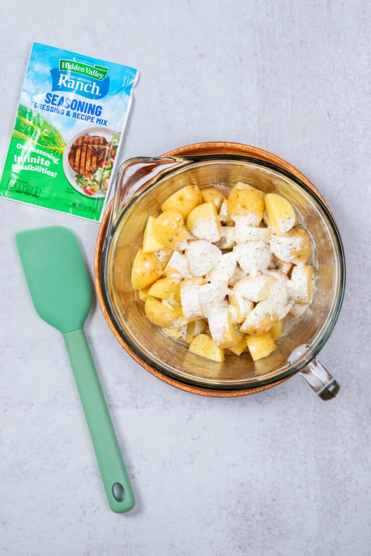 Sirring ranch seasoning mix to potatoes that are in a bowl. Next to the bowl is a packet of Hidden Valley Ranch seasoning mix.