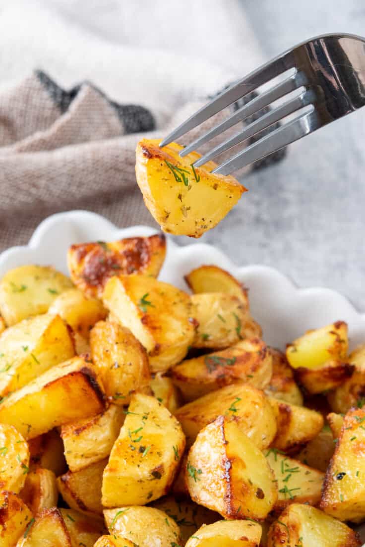 A roasted new potato seasoned with ranch and dill on a fork showing how soft and it is on the outside, and crispy on the outside. The fork is over a side dish.